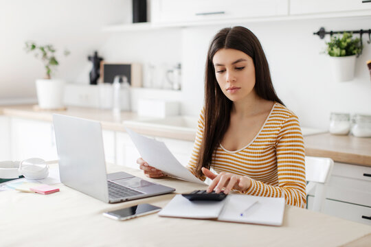 Lady doing paperwork and using calculator, accounting taxes and planning budget, sitting at table in kitchen and checking financial documents
