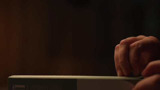 Close-up side view of unrecognizable woman carefully unboxing box with new electronic device in dark room on black background under soft illumination with warm light. v