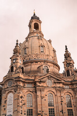 Fototapeta premium The Frauenkirche in Dresden, Germany, stands tall against a cloudy sky, framed by lush green leaves. The historic Baroque architecture contrasts with modern buildings in the foreground.