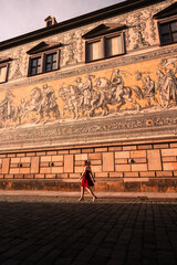 Obraz premium A woman in a red dress walks along the Fürstenzug mural in Dresden, Germany, during golden hour. The warm sunlight highlights the historic architecture and cobblestone street, creating a cinematic atm