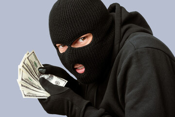 Closeup portrait of sneaky burglar with black mask and gloves counting money and looking at camera,...