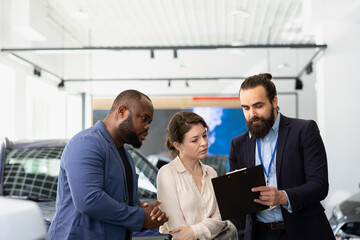 Car dealership salesman meeting with customers to discuss their needs, promoting sales offers and participating in test drives with interested customers, showing them offers on clipboard