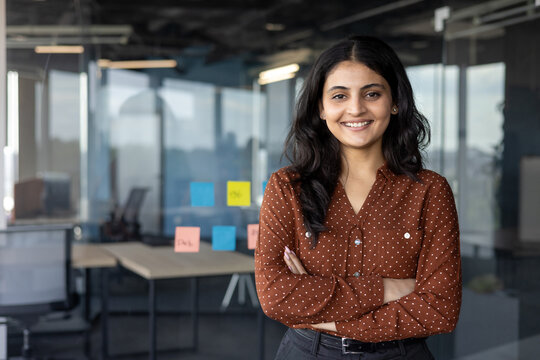 Successful confident businesswoman at workplace inside office, woman with crossed arms smiling and looking at camera, standing near window. Satisfied office worker of company.