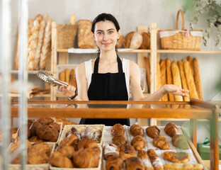 Young female salesperson at showcase with fresh pastries in bakery