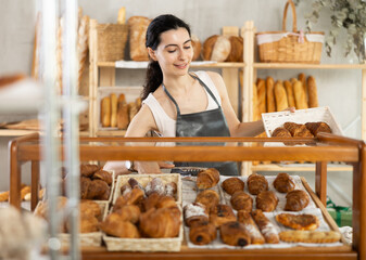 Authors bakery, family business idea. Young female sales consultant puts croissants on counter, complements assortment with fresh rolls, puts ready-made croissants in glass case