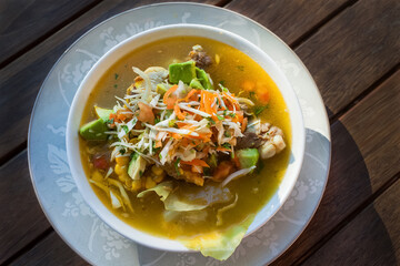 Ajiaco soup garnished with avocado, chopped vegetables, and chicken on wooden table