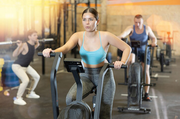 Young dedicated active strong fit woman doing cardio workout on stationary bicycle as hard...