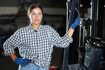 Young female worker in uniform posing by forklift in metallurgical workshop
