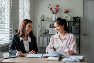 Two women in a modern office discussing work, surrounded by documents and laptops, showcasing teamwork and collaboration.