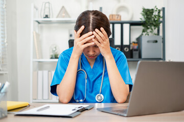 A healthcare worker in blue scrubs appears stressed while sitting at a desk with a laptop in a...