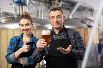 Portrait of positive man and woman brewmasters presenting beer in brewery.