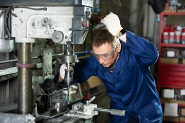 Experienced middle-aged machinist in blue work coat, protective gloves and glasses, focusing on metalworking in industrial workshop, confidently using drill press to create holes in workpiece..