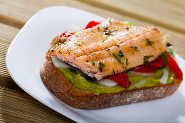 Healthy toast with baked trout fillet, guacamole, bell pepper and greens served on white plate