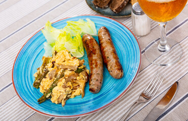 Scrambled eggs with mushrooms, chicken fillet and asparagus are served on plate. Fried pork sausages add richness to dish.