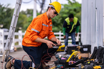 A skilled technician in a bright orange safety jacket and hard hat prepares electrical wiring with precision. Tools and open cases are spread in the background, showcasing a professional workspace.