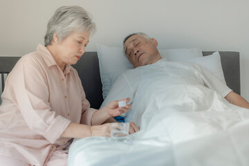 An elderly woman tends to her husband by administering medication while he rests in bed. The image...
