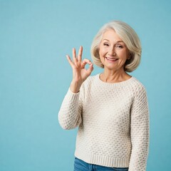 Obraz premium Cheerful smiling mature old woman lady in pastel white knitted sweater casual clothes standing showing Ok okay gesture sign looking camera isolated on blue light color background studio portrait 