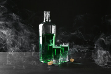 Absinthe in shot glasses, bottle and brown sugar on table against black background with smoke
