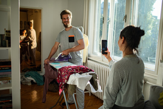 Woman taking picture of boyfriend ironing clothes at home
