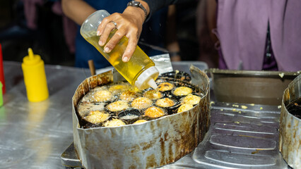 Street food vendor frying Takoyaki-style snacks in a round cast iron pan at a night market. A hand pours oil over golden-brown balls, sizzling in hot oil. Authentic street food scene in Asia.