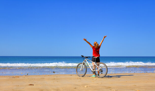 A woman stands on a sandy beach with her bicycle, arms raised towards the blue sky, celebrating freedom and adventure as ocean waves roll onto the shore