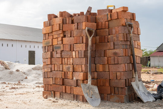 A large stack of red clay bricks neatly arranged at a construction site with two shovels leaning against them. The background features a white building and piles of sand.