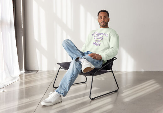 Mockup of man dressed in a light sweater and jeans sits confidently on a chair, looking directly at the camera in a stylish interior.