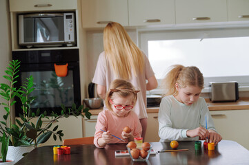 Two daughters paint eggs at table while mother works in background, creating quiet and focused Easter preparation scene