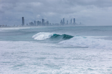 A large windswept wave, part of the effects of the edge of cyclone Alfred off the Queensland coast, breaks with Surfers Paradise in the background under threatening skies on the Gold Coast, Australia.