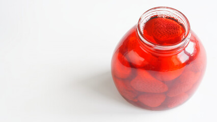 Refreshing homemade red strawberry preserves in a open glass jar on a clean white surface with fresh fruit and syrup. Copy space. Photo