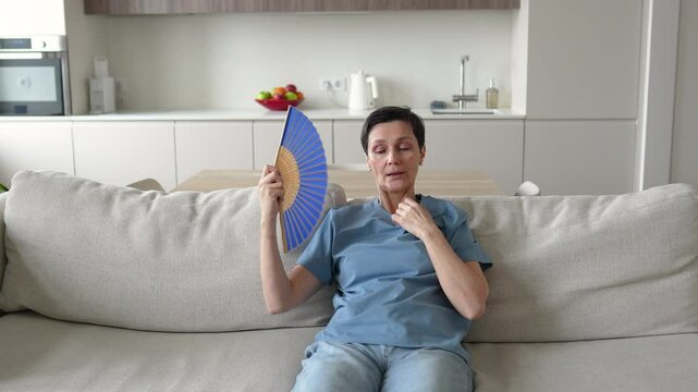 Unwell exhausted mature woman sitting on home couch waving paper fan to cool down. Overheated annoyed female suffers from stuffiness, feeling hot during heatwave in apartment without air conditioning.