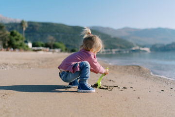 Little girl digs sand on the beach with a toy shovel. Side view