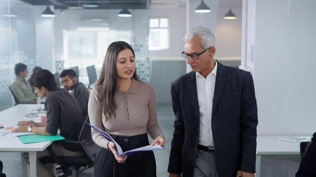 A senior corporate businessman or aged executive walking and collaborating on a project with a woman office employee, with coworkers or colleagues working in a modern start up business workplace.