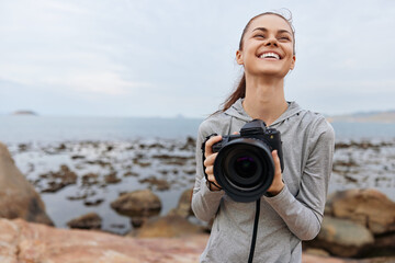 Happy woman holding a camera enjoys nature on a rocky beach, wearing a gray hoodie, with an emotional smile and a soft ocean background