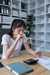 Young woman analyzing receipt and financial documents at home office while experiencing confusion and concern