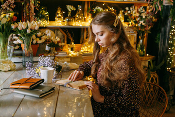 Young woman with long wavy hair reading a book at a table surrounded by flowers and warm fairy lights. Calm and tranquil indoor scene highlighting a love for literature and nature.