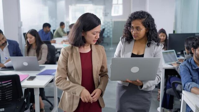 Two modern female corporate friendly associates walking and using a laptop to discuss an assignment together, office employees are busy working in the background in a modern diverse business workplace