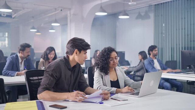 Two modern Indian Asian male and female coworkers team up or collaborate together on an assignment using a laptop, office employees are busy working in the background in a modern start up business.