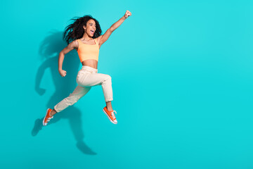 Energetic woman jumping joyfully wearing casual fashion against a vibrant teal background