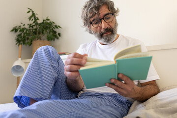 Mature man wearing pajamas reading a book relaxing in bed.