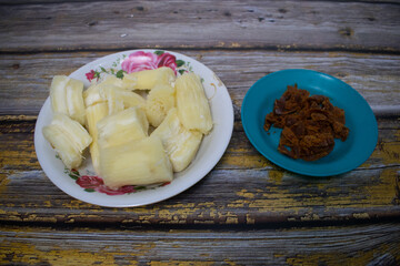A plate of boiled cassava cut into pieces is served on a floral patterned plate, accompanied by brown sugar
