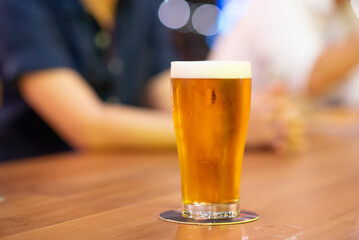 Beer in the long glass on the table at night bar with background people meeting in the club. Hang out concept.