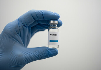 Peptide vial held in gloved hand. Close-up of a gloved hand holding a vial labeled "Peptide". The vial contains a clear liquid and is capped with a silver top