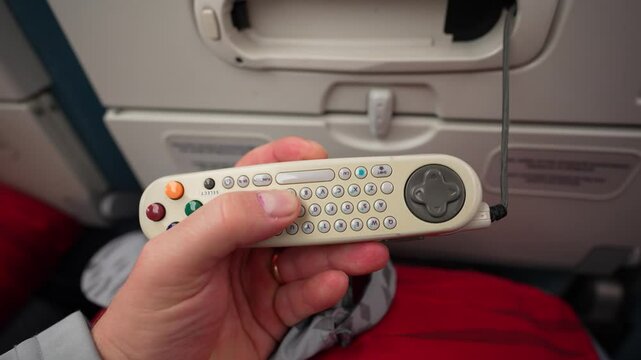 A passenger holds an in-flight entertainment system remote control with buttons and a joystick, connected to the airplane seat.