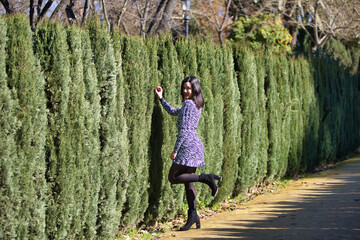 Portrait of a young, beautiful Latin brunette woman in front of the green hedges separating a road....