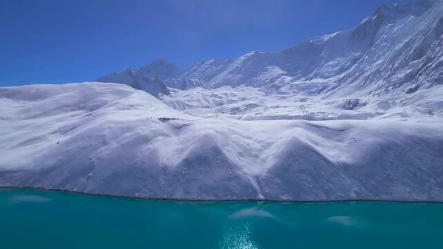 Aerial view of blue Tilicho Lake in Nepal. Drone slowly moves towards to lake shore and white snow-covered rugged landscape, snowy jagged mountain peaks in the horizon. Clear blue sky and sunny day