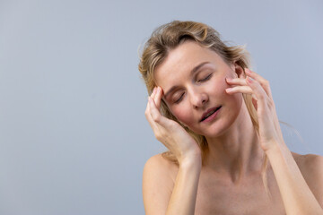 Fototapeta premium Woman with Light Hair Using Hands to Ease Stress on the Face in a Relaxed Expression Against a Soft Gray Background, Emphasizing Comfort and Serenity in a Calm Setting