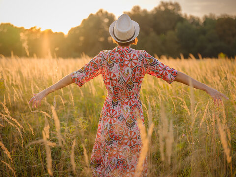 Young woman walking through picturesque European field in late summer. Golden sunlight, lush greenery, and serene rural atmosphere create peaceful countryside scene. - Powered by Adobe