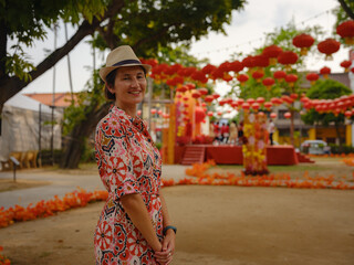 Fototapeta premium Young woman in ethnic dress and hat exploring festive streets of George Town, Malaysia, during Chinese New Year. Vibrant lanterns, cultural celebrations, and historic charm create a unique atmosphere