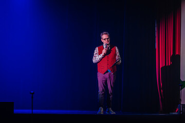 Fototapeta premium A presenter in a colorful outfit speaks into a microphone on stage under dramatic lighting, holding a paper. A red curtain is in the background..